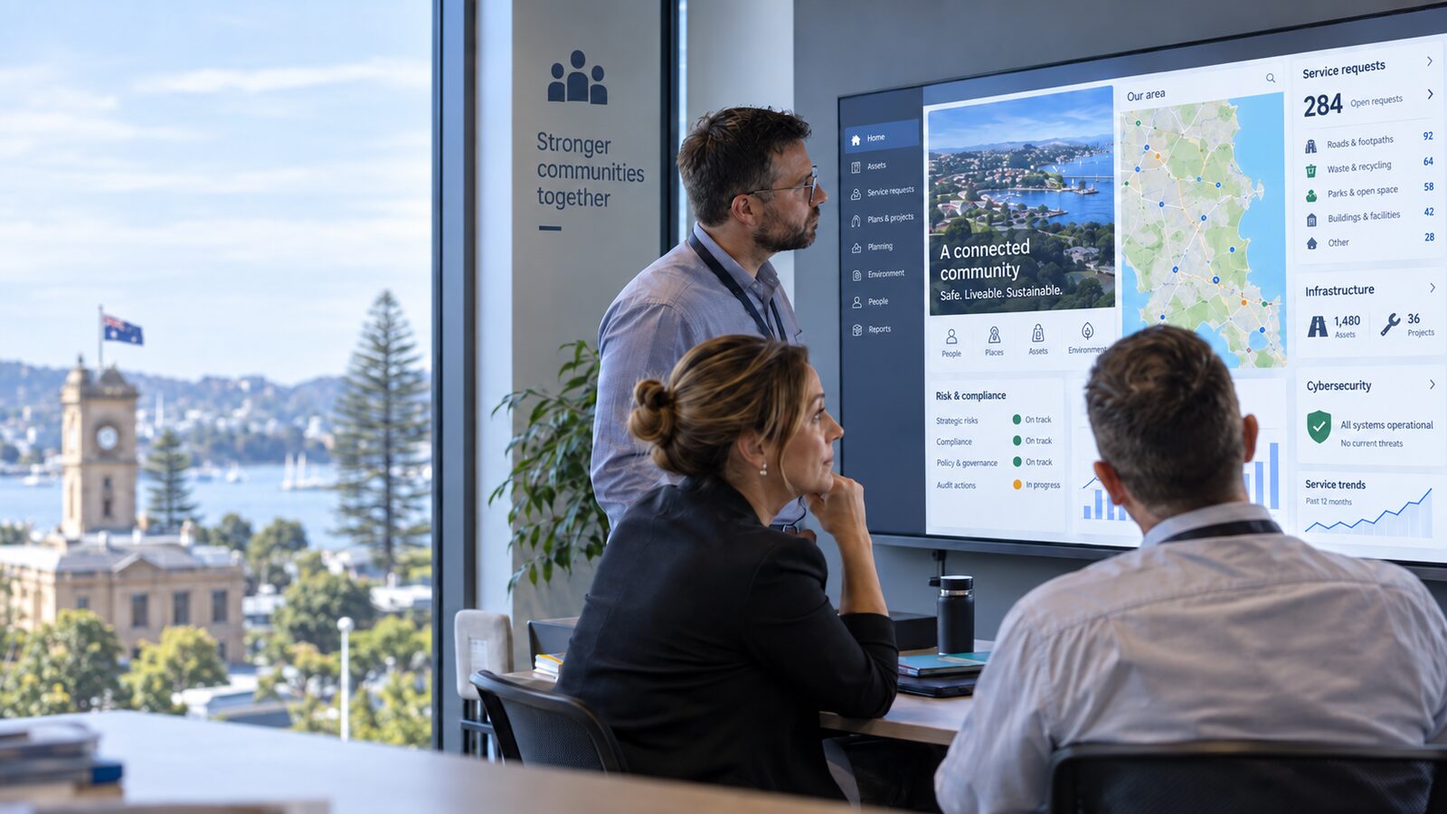 Council staff reviewing a community services, infrastructure and cybersecurity dashboard, with a coastal town and town hall visible through the window.